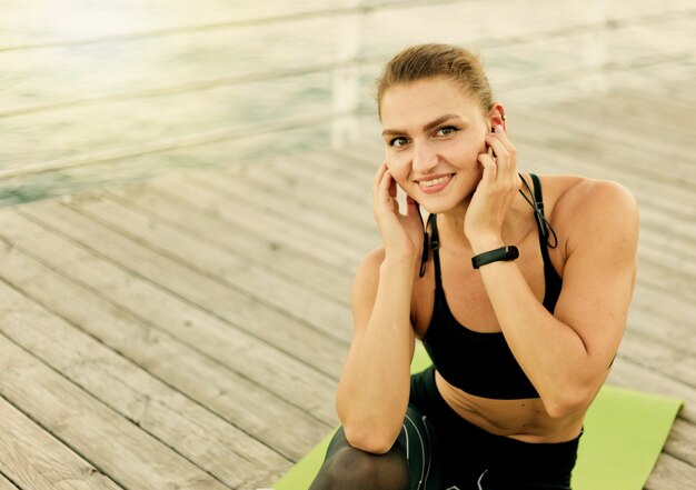 Jeune femme fit écouter de la musique avec des écouteurs alors qu'il était assis sur un tapis à une terrasse en bois sur la plage