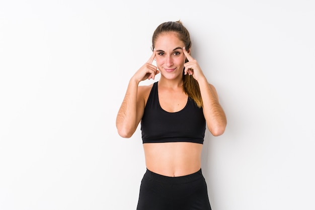 Jeune femme fitness caucasien posant dans un mur blanc concentré sur une tâche, gardant les index pointant la tête.