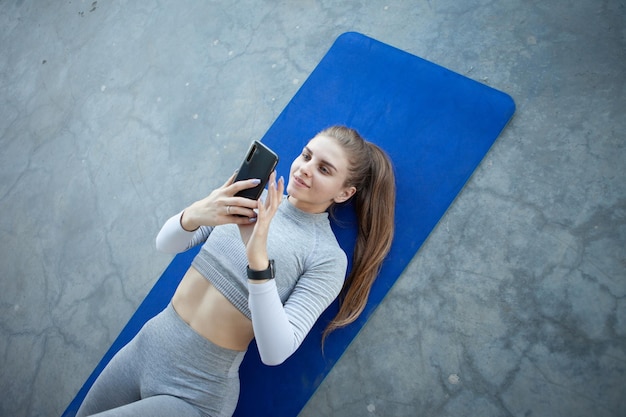 Une jeune femme de fitness caucasienne se trouve sur un tapis et utilise un smartphone sur un sol en béton