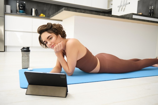 Jeune femme fitness fille en survêtement allongé sur un tapis de fitness en regardant le didacticiel vidéo yoga pilates sur