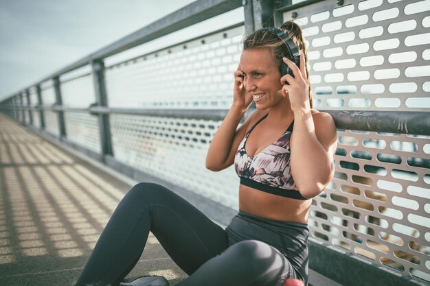 Une jeune femme de fitness se repose après un entraînement intensif sur le pont de la rivière et met ses écouteurs.
