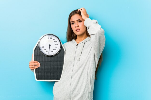 Jeune femme de fitness tenant une balance choquée, elle se souvient d'une réunion importante.