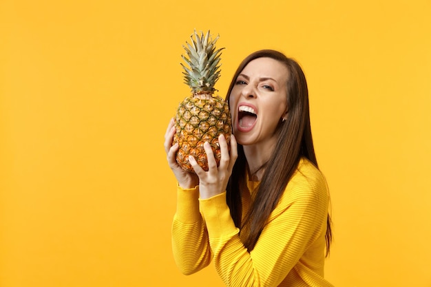 Jeune femme folle dans des vêtements décontractés tenant, mordant des fruits d'ananas mûrs frais isolés sur fond de mur orange jaune en studio. Mode de vie vivant des gens, concept de vacances relaxantes. Maquette d'espace de copie