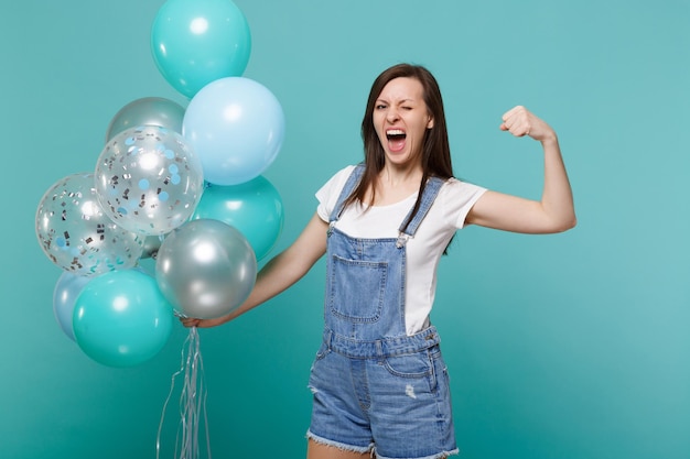 Jeune femme folle qui crie et qui clignote, montrant des biceps, des muscles célébrant, tenant des ballons à air colorés isolés sur fond bleu turquoise. Fête d'anniversaire, concept d'émotions de personnes.