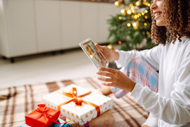 Une jeune femme sur fond d'arbre de Noël avec des cadeaux avec tablette a un appel vidéo ou un chat vidéo