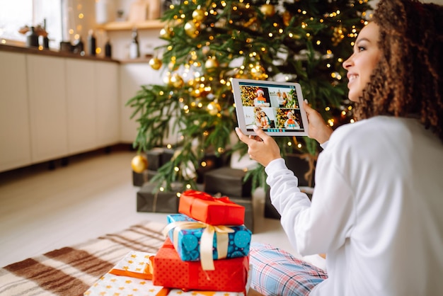 Une jeune femme sur fond d'arbre de Noël avec des cadeaux avec tablette a un appel vidéo ou un chat vidéo