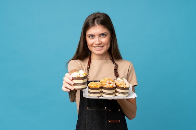 Jeune femme sur fond bleu isolé, tenant des mini gâteaux et l'offrant