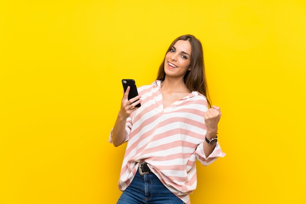 Jeune femme sur fond jaune isolé avec téléphone en position de victoire