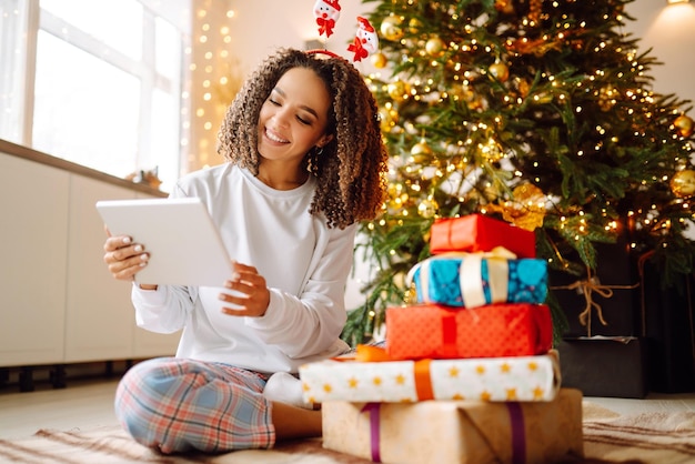 Une jeune femme sur le fond d'un sapin de Noël avec des cadeaux avec une tablette passe un appel vidéo.