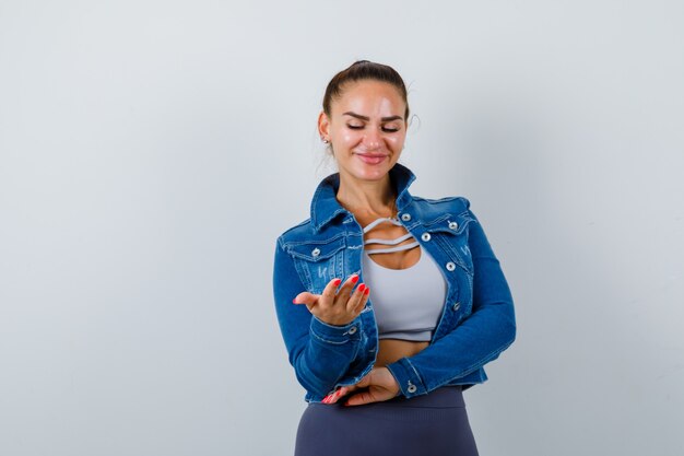 Jeune femme en forme en haut, veste en jean regardant la paume étalée et l'air heureux, vue de face.