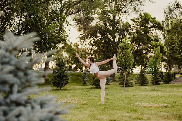 Jeune femme en forme pratique le yoga dans un parc verdoyant sur l'herbe le matin d'été Tenez-vous dans la pose de la danseuse en vêtements de sport équilibrant l'exercice fond de la forêt Concept de mode de vie sain et d'harmonie avec le corps