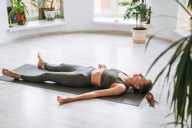Jeune femme en forme pratique le yoga faisant du savasana dans un studio de yoga léger avec une plante verte
