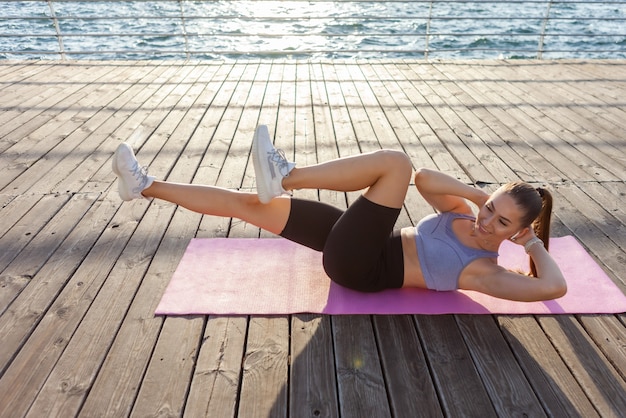 Une jeune femme en forme en vêtements de sport entraîne les muscles abdominaux en position allongée sur le tapis au lever du soleil sur la plage