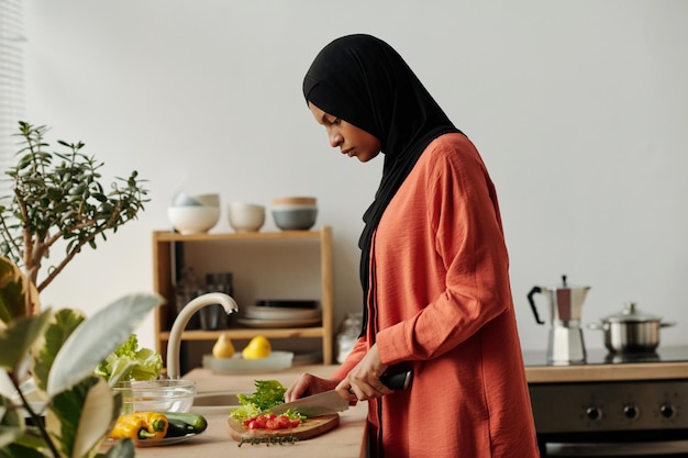 Une jeune femme avec un foulard noir et une chemise rouge coupe des légumes frais sur une planche à découper