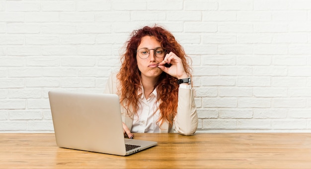 Jeune femme frisée rousse travaillant avec son ordinateur portable avec les doigts sur les lèvres, gardant un secret.