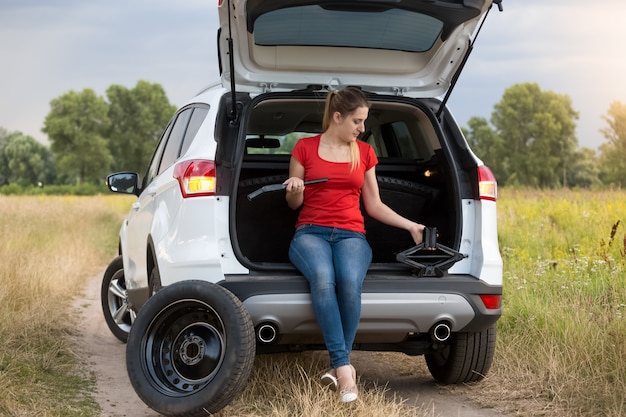 Jeune femme frustrée assise dans une voiture cassée regardant les outils pour changer les roues