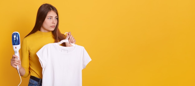 Jeune femme fumant ses vêtements à la maison, tenant un fer à vapeur et un t-shirt blanc sur des cintres dans les mains, regardant de côté avec un regard sérieux, posant contre le mur jaune.