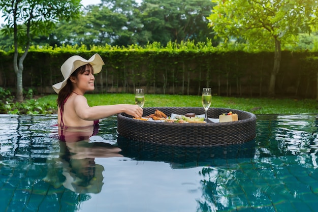 Jeune femme gaie appréciant avec la nourriture flottante et le verre de champagne dans la piscine