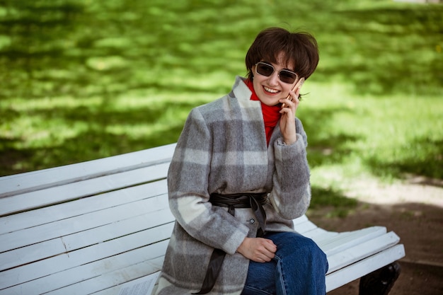Jeune femme gaie en manteau parlant au téléphone alors qu'il était assis sur un banc au printemps