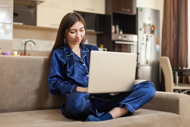 Jeune femme gaie en nuisette regarde dans l'écran de l'ordinateur portable tout en étant assise sur le canapé dans le salon