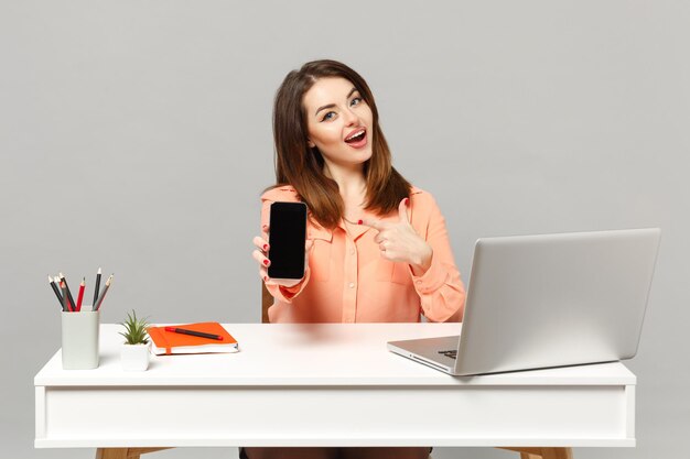 Jeune femme gaie pointant l'index sur un téléphone portable avec un écran vide vide assis au bureau avec un ordinateur portable pc isolé sur fond gris. Concept de carrière d'affaires de réalisation. Maquette de l'espace de copie.