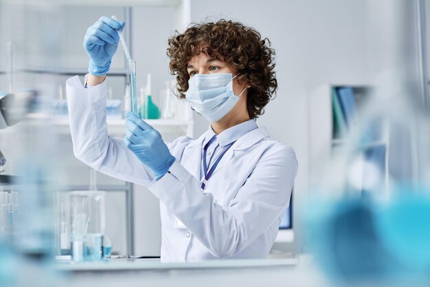 Jeune femme en gants de laboratoire et masque mélangeant deux substances liquides dans un flacon tout en créant un nouveau composé pour le vaccin en laboratoire