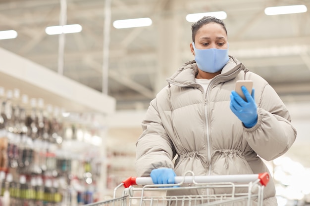 Jeune femme en gants de protection et masque lisant un message sur son téléphone mobile tout en faisant des achats avec panier en supermarché