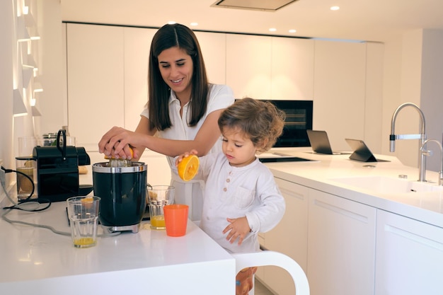 Une jeune femme et un garçon positives en vêtements décontractés faisant du jus d'orange à l'aide d'un extracteur de jus au comptoir de la cuisine dans un appartement moderne