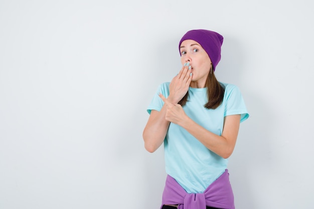 Jeune femme gardant la main sur la bouche, pointant vers le coin supérieur gauche en t-shirt, bonnet et l'air surpris. vue de face.