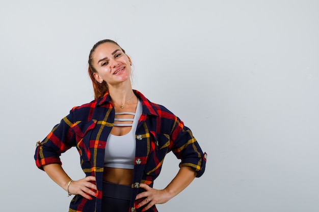 Jeune femme gardant les mains sur la hanche dans un haut court, une chemise à carreaux, un pantalon et l'air heureux, vue de face.