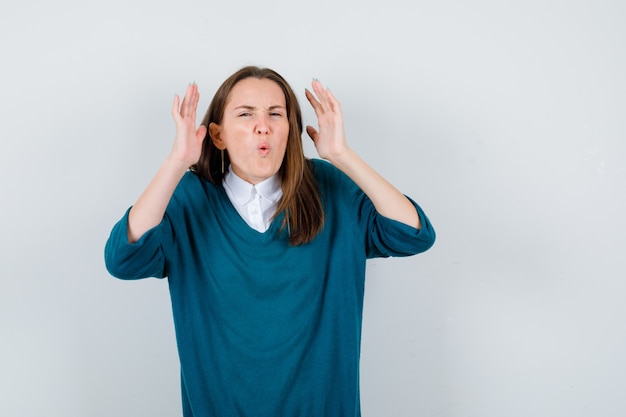 Photo jeune femme gardant les mains près de la tête en pull sur chemise blanche et semblant gênée. vue de face.