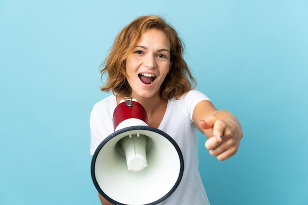 Jeune femme géorgienne isolée sur fond bleu criant à travers un mégaphone pour annoncer quelque chose tout en pointant vers l'avant