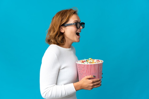 Jeune femme géorgienne isolée sur fond bleu avec des lunettes 3d et tenant un grand seau de pop-corn