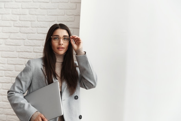 Jeune femme gestionnaire dans des verres debout près de la fenêtre du bureau, tenant un ordinateur portable à la main.