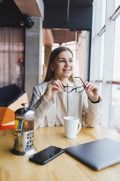 Une jeune femme gestionnaire dans une veste légère et des lunettes une fille avec un sourire sur son visage est assise à une table en bois avec un ordinateur portable et un téléphone Travail à distance