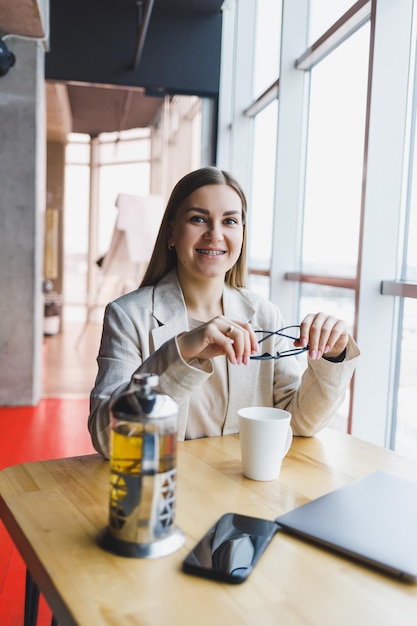 Une jeune femme gestionnaire dans une veste légère et des lunettes une fille avec un sourire sur son visage est assise à une table en bois avec un ordinateur portable et un téléphone Travail à distance