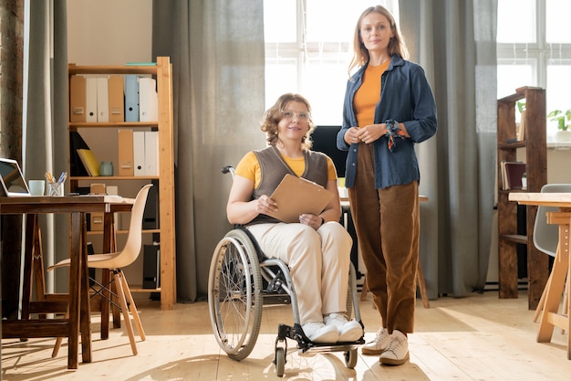 Jeune femme gestionnaire et sa collègue handicapée debout au centre du bureau entre les lieux de travail contre la fenêtre et les étagères avec des documents