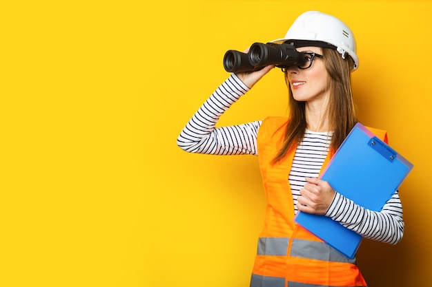 Jeune femme en gilet de signalisation et casque de construction à la recherche à travers des jumelles sur jaune