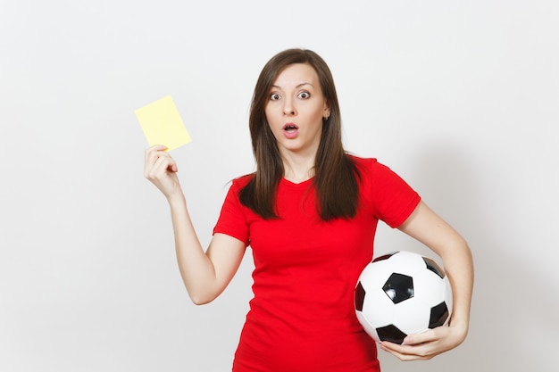 Jeune femme grave européenne, arbitre de football ou joueur en uniforme rouge montrant un carton jaune, tenant un ballon de football isolé sur fond blanc. Le sport joue au football, concept de mode de vie sain.