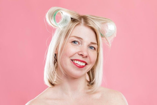 Jeune femme avec de gros bigoudis sur la tête Belle femme blonde avec des taches de rousseur se coiffe Beauté et soins personnels Gros plan Fond rose