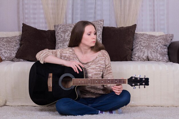 Jeune femme avec une guitare acoustique noire sur un tapis dans la chambre