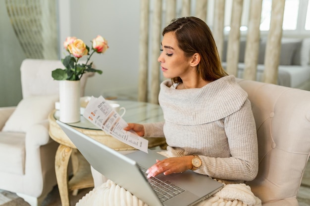 Jeune femme habillée avec désinvolture tenant des papiers dans ses mains, calculant et payant des factures à la maison. Heureuse belle femme assise sur le canapé avec ordinateur portable calculant les finances de la maison. Travail à domicile