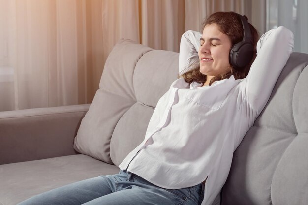 Jeune femme handicapée aux cheveux longs atteinte de paralysie cérébrale se détend en souriant et en écoutant de la musique dans des écouteurs en s'appuyant sur un canapé à la lumière du soleil à la maison