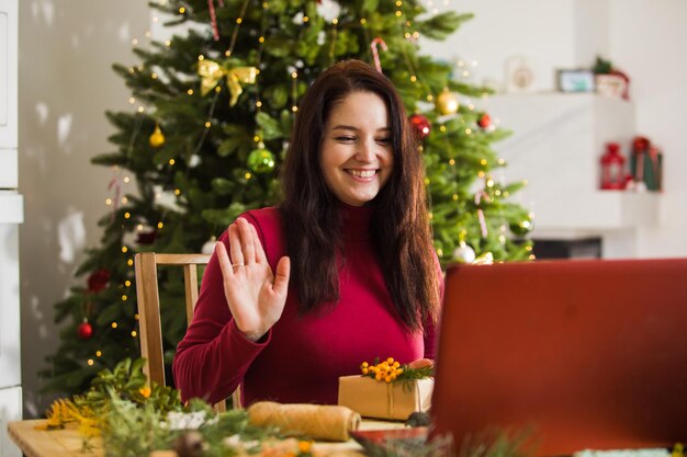 La jeune femme heureuse agite la main à la caméra de l'ordinateur portable Femme assise devant un ordinateur portable sur le fond de l'intérieur de Noël