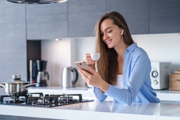 Jeune femme heureuse à l'aide d'un smartphone et d'un casque sans fil pour lire un livre audio pendant la pause-café dans la cuisine à la maison. Personnes mobiles modernes