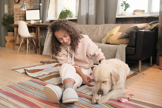 Jeune femme heureuse assise sur le sol et jouant avec son animal de compagnie dans la chambre