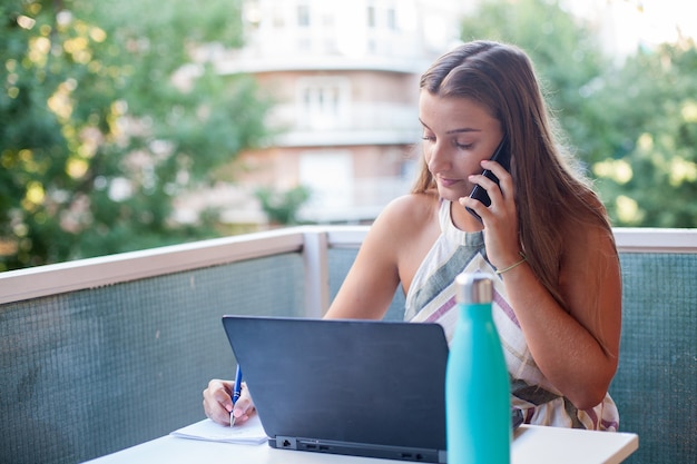 Jeune femme heureuse assise sur une terrasse ayant un appel avec son téléphone portable, travaillant sur ordinateur à la maison