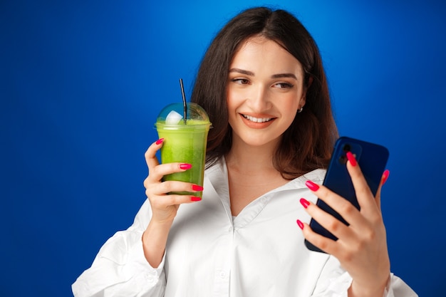 Jeune femme heureuse attirante tenant une tasse de smoothie vert et utilisant son smartphone contre b bleu...