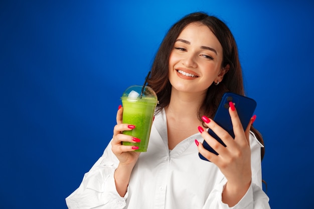 Jeune femme heureuse attirante tenant une tasse de smoothie vert et utilisant son smartphone contre b bleu...