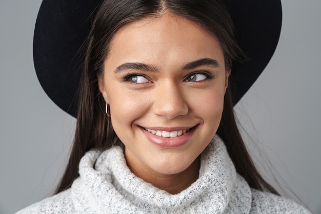 jeune femme heureuse au chapeau noir souriant et regardant de côté isolé sur un mur gris